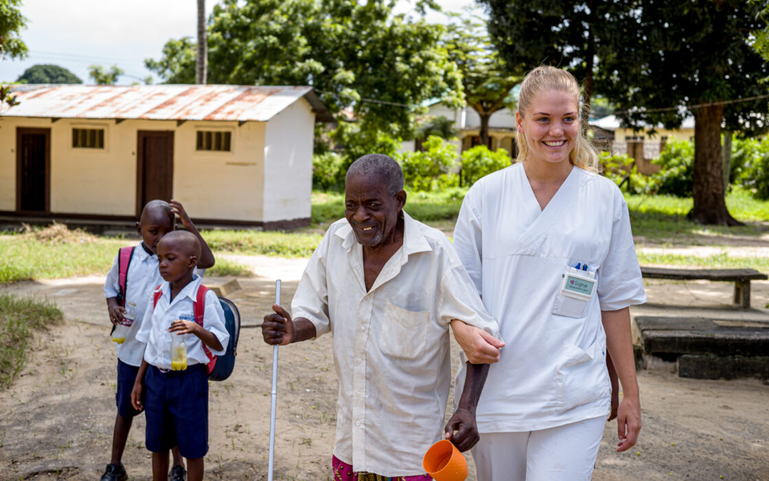 Student supporting an elderly man with walking, with children in the background