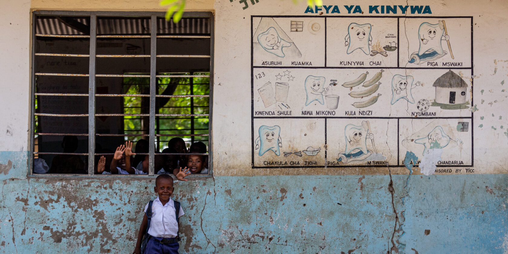 Child smiling while standing in front of a classroom window with other children waving through it